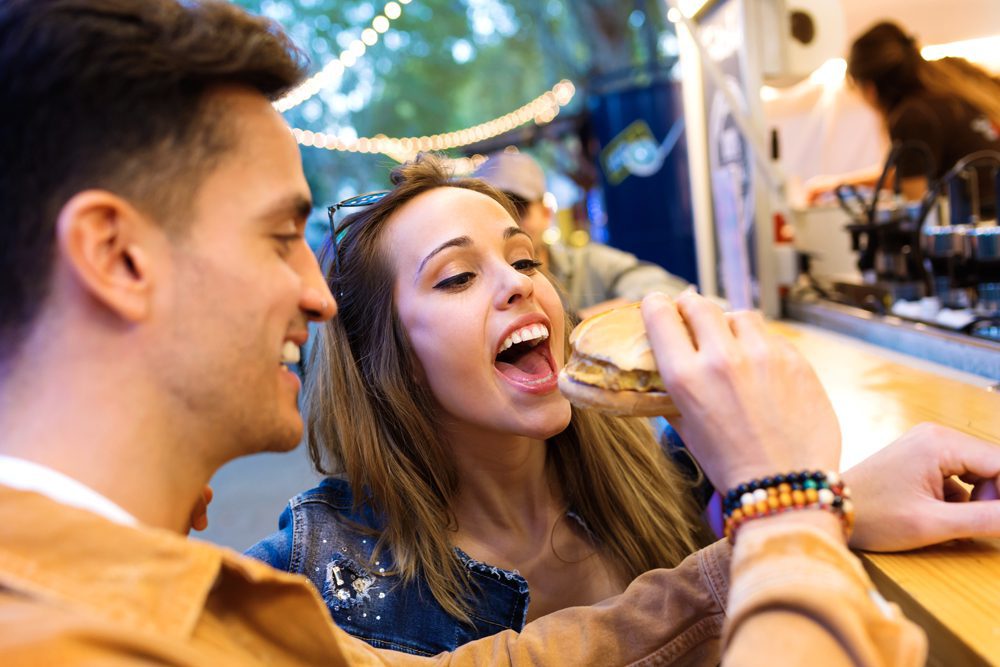 Attractive young man giving hamburger to his beautiful girlfriend in eat market in the street.